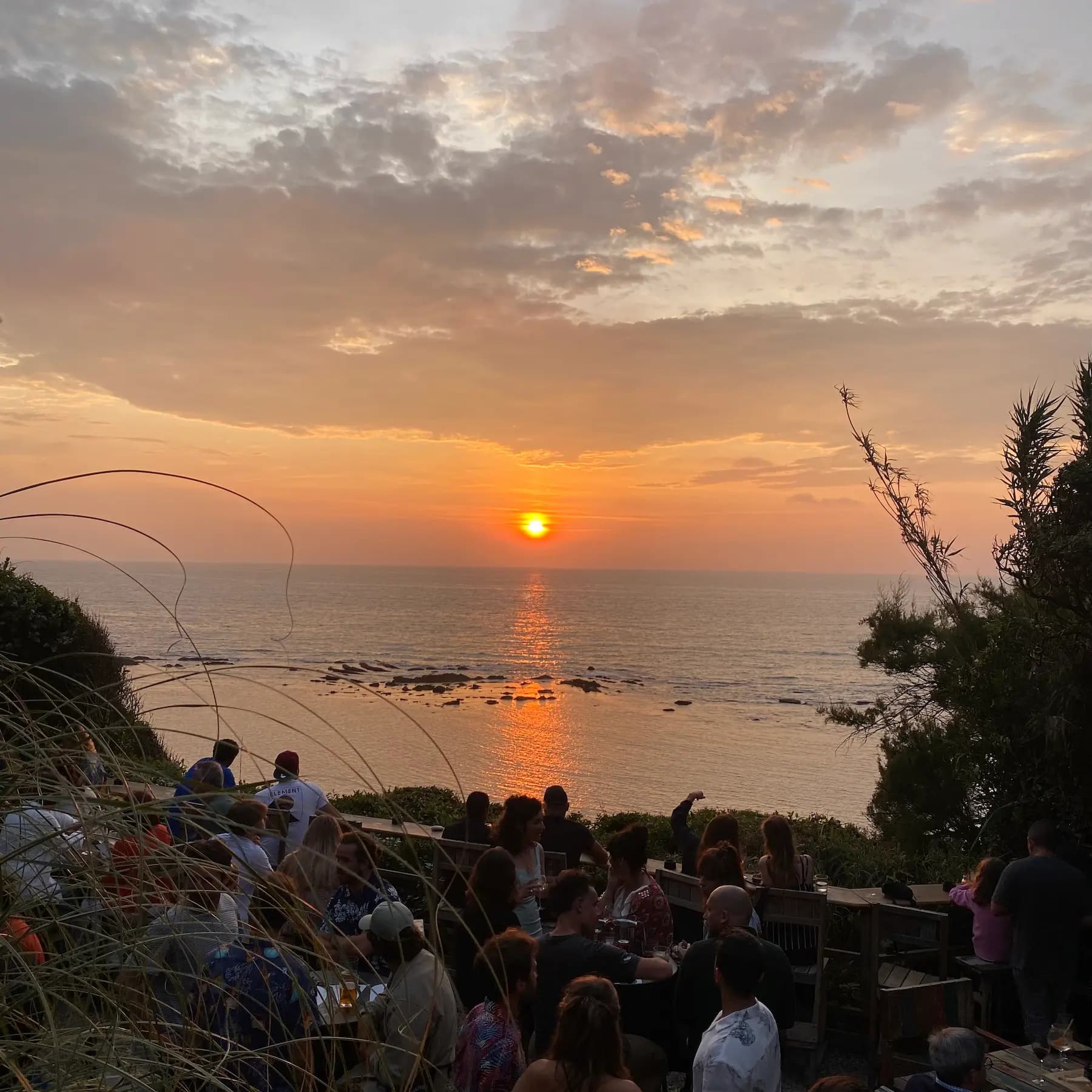 A crowd gathered at an open-air clifftop bar watching the sun set over the ocean